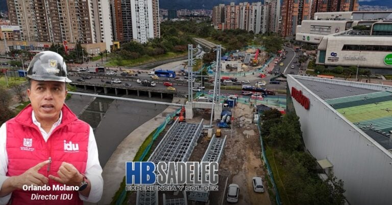 Spokesperson Orlando Molano in a red IDU vest and hard hat speaks in the foreground as a construction site and busy city street stretch behind him, with HBSADELEC branding visible. - Las2orillas.co: Historias, voces y noticias de Colombia