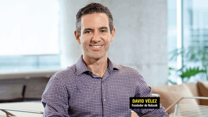 Smiling man in a purple checkered shirt seated in a bright, modern office; a yellow-and-black badge on his chest reads 'Founder of Nubank' (logo not stated). - Cuál será el cargo de David Vélez, el cerebro de Nubank, en el gobierno De la Espriella