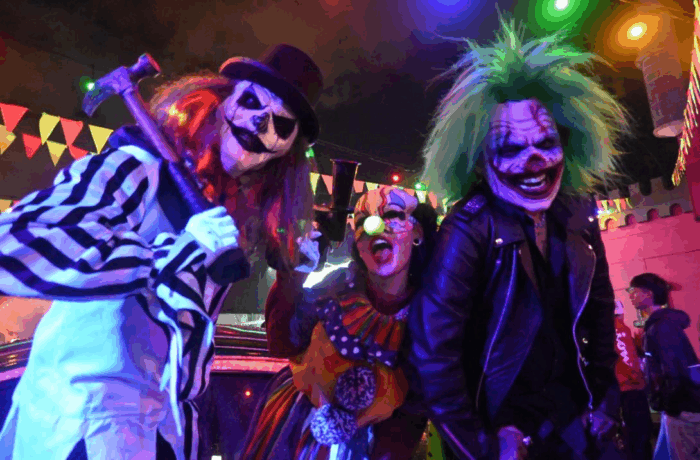 Los fiscales que lograron la condena por el asesinato del estudiante de los Andes ocurrido el pasado Halloween Three Halloween-costumed performers under colorful lights: a striped prisoner-clown with a top hat and an axe prop, a rainbow clown, and a green-haired rocker in a black leather jacket posing for the camera. - Los fiscales que lograron la condena por el asesinato del estudiante de los Andes ocurrido el pasado Halloween