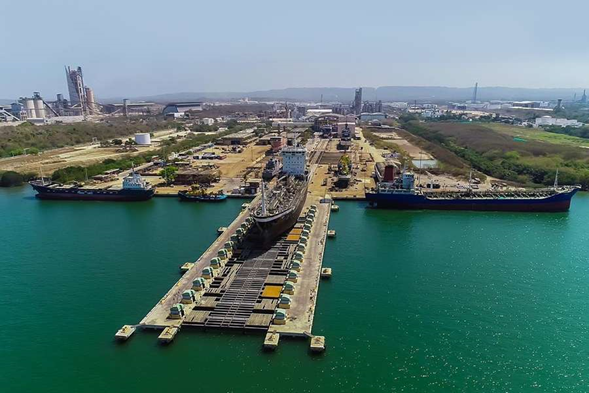 Colombia se mete en las grandes ligas navales y en Cartagena construye su primer buque de guerra de ,6 billones Aerial view of a harbor shipyard with a long dry dock and a ship under construction, flanked by two large cargo ships on turquoise water. - Colombia se mete en las grandes ligas navales y en Cartagena construye su primer buque de guerra de ,6 billones