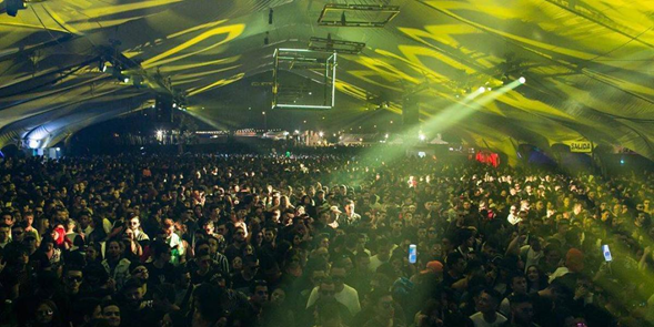 En una casa clandestina en La Macarena nació Baum, el festival de electrónica que mueve más de .000 millones Dense crowd at a large indoor concert tent, with yellow-green stage lighting sweeping over the audience and a distant stage. - En una casa clandestina en La Macarena nació Baum, el festival de electrónica que mueve más de .000 millones