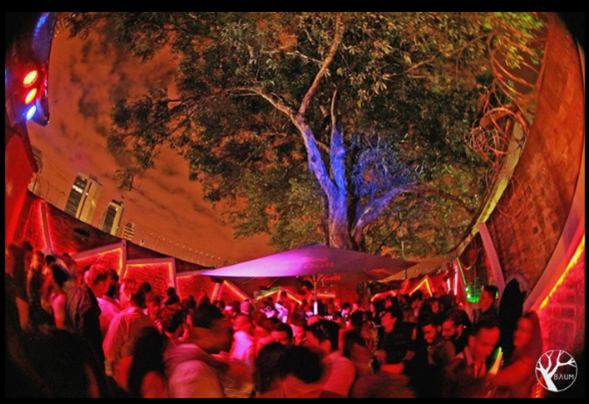 En una casa clandestina en La Macarena nació Baum, el festival de electrónica que mueve más de .000 millones Crowd of people at an outdoor nighttime party with colored lights, a central tree, and a large umbrella. - En una casa clandestina en La Macarena nació Baum, el festival de electrónica que mueve más de .000 millones