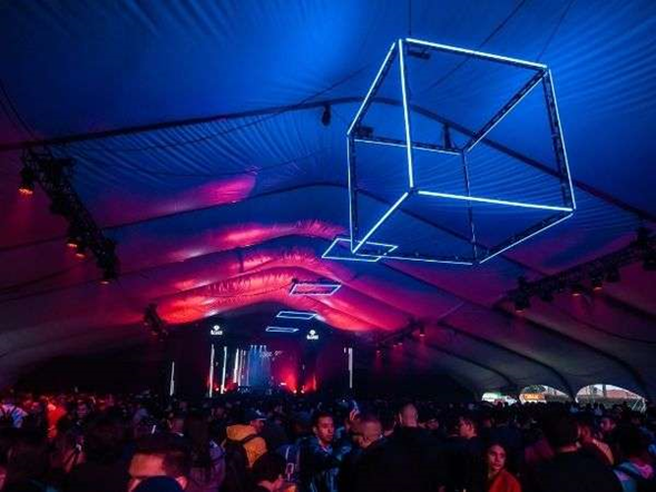 En una casa clandestina en La Macarena nació Baum, el festival de electrónica que mueve más de .000 millones Neon cube sculpture hanging from a tent ceiling over a crowded event space with colorful blue and pink lighting. - En una casa clandestina en La Macarena nació Baum, el festival de electrónica que mueve más de .000 millones