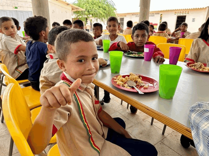 El fantasma del zar del PAE sigue rondando y hoy la Contraloría tiene mil millones bajo la lupa Boy at a school lunch table gives a thumbs-up to the camera, surrounded by classmates eating on colorful plastic plates and cups in a bright cafeteria. - El fantasma del zar del PAE sigue rondando y hoy la Contraloría tiene mil millones bajo la lupa