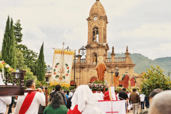 Baloto cayó en un pequeño pueblo de Boyacá, el departamento donde hace diez años no caía el premio gordo Religious procession on a street toward a stone church with statues and floral tributes; banner reads JHS. - Baloto cayó en un pequeño pueblo de Boyacá, el departamento donde hace diez años no caía el premio gordo