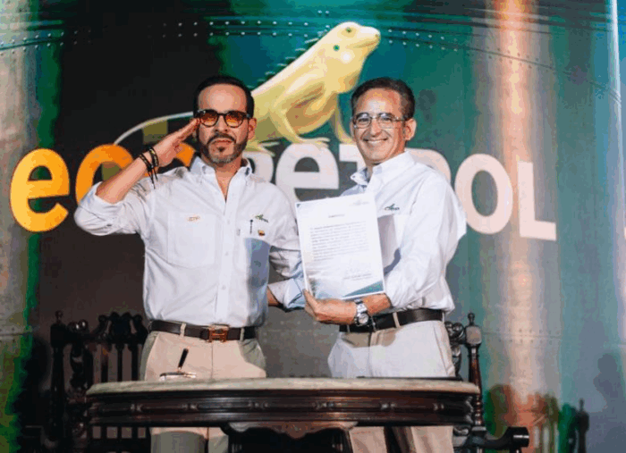 El remezón en Ecopetrol con el que el nuevo presidente Joaquín Gutiérrez arma su equipo de vicepresidentes Two men in white shirts pose on stage; one salutes while the other holds a document, with a frog image and green backdrop behind. - El remezón en Ecopetrol con el que el nuevo presidente Joaquín Gutiérrez arma su equipo de vicepresidentes