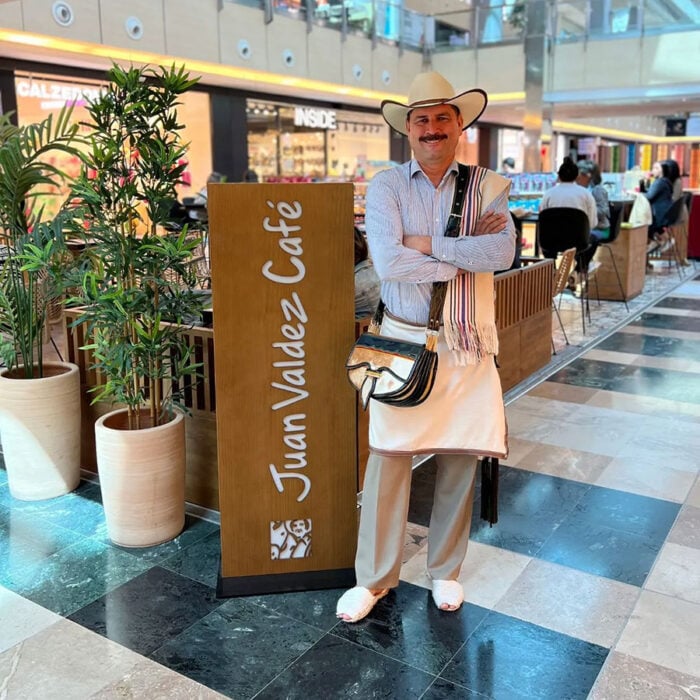 La jugada de Omar González con el grupo Trinity para llevar las tiendas Juan Valdez a Cataluña: abrirá diez en Barcelona Man in a cowboy outfit posing with arms crossed beside a Juan Valdez Café sign in a shopping mall.