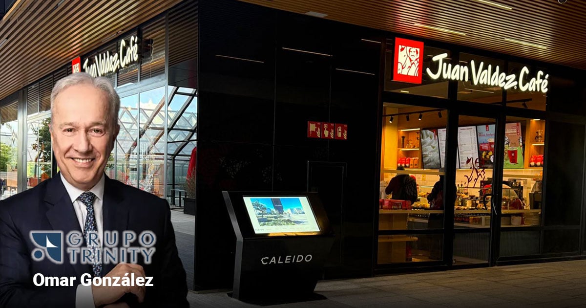 Smiling man in a suit stands outside a Juan Valdez Café storefront with a touchscreen information kiosk nearby and Grupo Trinity branding visible. - La jugada de Omar González con el grupo Trinity para llevar las tiendas Juan Valdez a Cataluña: abrirá diez en Barcelona
