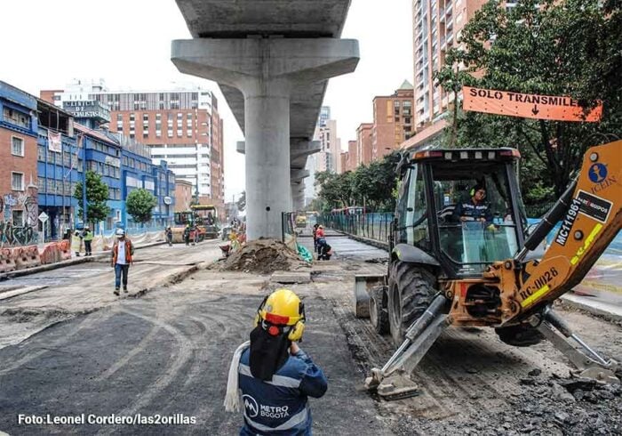 Luz verde del Tigre para que los bogotanos empiecen a soñar con la Línea 4 del metro Construction crew works on a city street beneath an elevated rail structure, with a yellow backhoe loader and workers in hard hats nearby. - Luz verde del Tigre para que los bogotanos empiecen a soñar con la Línea 4 del metro