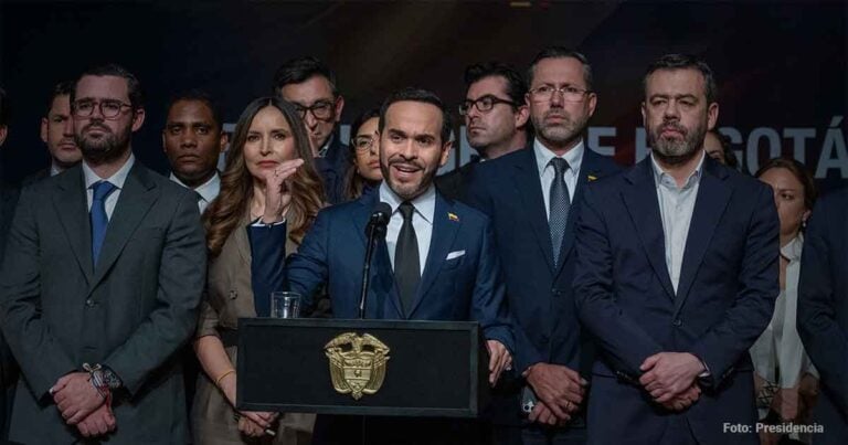 Man in blue suit speaking at a lectern with a crest, flanked by delegates in business attire behind him at a formal event. - Las2orillas.co: Historias, voces y noticias de Colombia