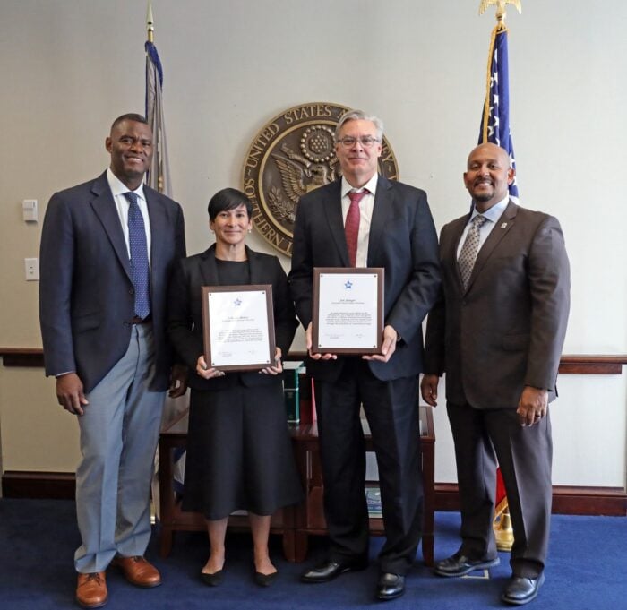 Five professionals in business attire pose for a photo in a formal office, two holding framed certificates, with a large government seal and flags in the background. - La fuerte fiscal de Miami con sangre colombiana que está detrás de los casos de Álex Saab y su socio Álvaro Pulido