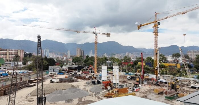El megaedificio de 23 pisos y medio billón de pesos que la Alcaldía de Bogotá levanta cerca de Corferias Active construction site with two tall yellow cranes over machinery and piles of gravel against an urban skyline and mountains in the distance. - El megaedificio de 23 pisos y medio billón de pesos que la Alcaldía de Bogotá levanta cerca de Corferias