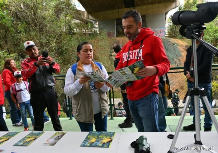 El sendero turístico que lleva a las montañas de Bogotá al que Galán le metió 0 millones para recuperarlo Two adults read brochures at an outdoor info table while a camera on a tripod records nearby. - El sendero turístico que lleva a las montañas de Bogotá al que Galán le metió 0 millones para recuperarlo